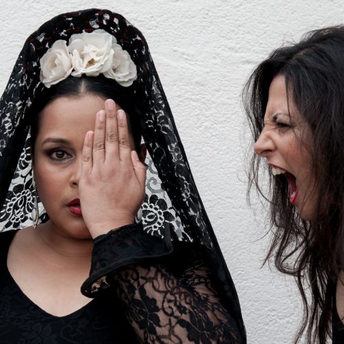 A Flamenco dancer dressed in black, medium close-up and facing camera covers her left eye as another dancer appears to be yelling into her left ear.