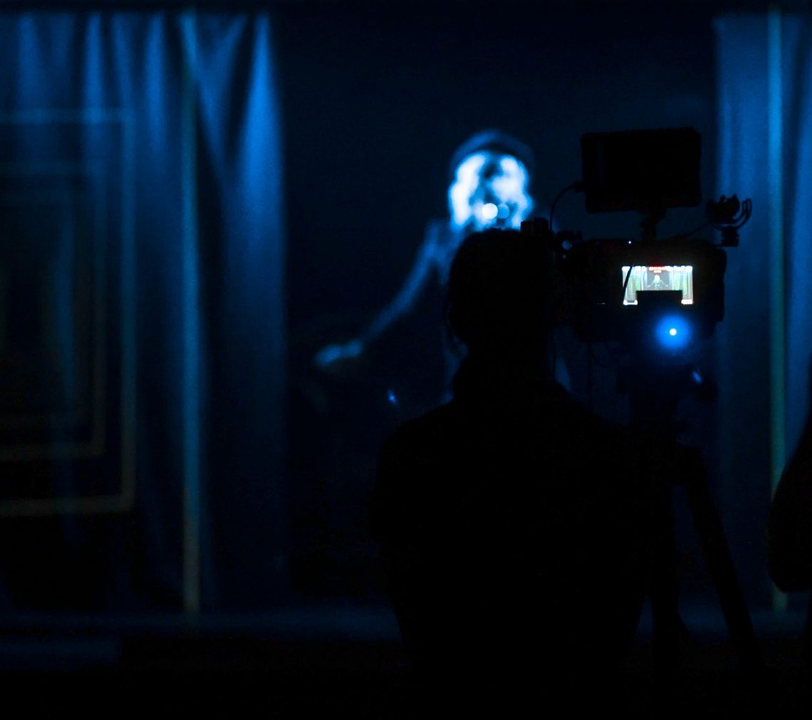 A darkened stage where a cabaret performer is about to hit the stage. Behind her is the screen of a digital camera, recording her every move.